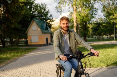 Genç bir adam bisikletini dolambaçlı bir patika boyunca ağaçlarla ve açık alanlarla dolu sakin bir parkta sürüyor. Sıcak öğleden sonra ışığı ve temiz havanın tadını çıkarıyor..