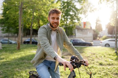 Genç bir adam yeşil bir parkta bisiklet sürüyor, keyifli havanın tadını çıkarırken gülümsüyor. Uzun ağaçlar arkaplanı kentsel elementlerle çerçeveletir.