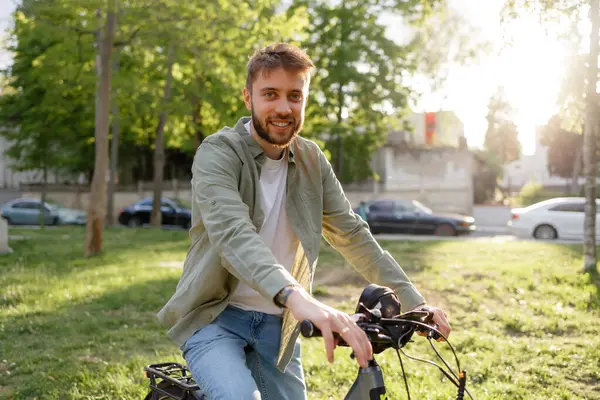 Genç bir adam yeşil bir parkta bisiklet sürüyor, keyifli havanın tadını çıkarırken gülümsüyor. Uzun ağaçlar arkaplanı kentsel elementlerle çerçeveletir.
