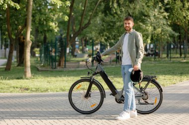 Genç bir adam parkta elektrikli bisikletinin yanında dururken gülümsüyor. Bir kask tutuyor ve günlük kıyafetler giyiyor. Yeşillikler arasındaki hoş havanın tadını çıkarıyor..