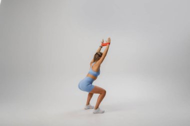 Parlak, ferah bir stüdyoda spor kıyafetleri giyerken kolları havada çömelir. Formuna odaklanıldığında, dayanıklılık eğitimi ve egzersiz azmi vurgulanıyor..