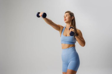 Bir kadın antreman yapıyor, açık mavi iç çamaşırı giyerken halterleri kafasının üstüne kaldırıyor. Arka plan çok az, egzersizi için odaklanmış bir atmosfer yaratıyor..