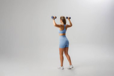 Bir kadın arkası seyirciye dönük duruyor, mavi spor kıyafetlerini giyerken dambılları kaldırıyor..