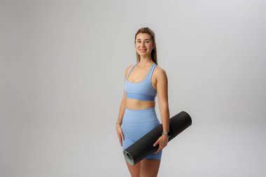 Hafif bir stüdyoda elinde yoga minderiyle gülümseyen bir kadın var. Rahat idman kıyafetleri içinde yoga ya da fitness seanslarına hazır olduğunu gösteriyor..