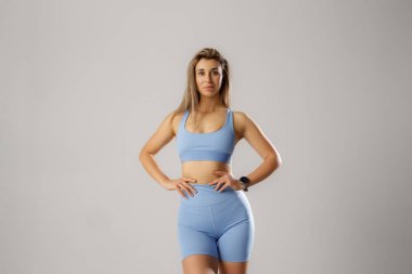 Genç bir kadın rahat bir poz veriyor, açık mavi spor kıyafetini tarafsız bir arkaplanda sergiliyor. Kameraya gülümserken kendine güveni tam bir güç anı yakalıyor..