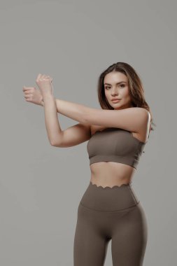 Bir kadın şık bir spor kıyafeti giyerken kollarını esnetiyor. Ayar, fitness temalı bir oturum için mükemmel bir minimalist gri arkaplan içeriyor.