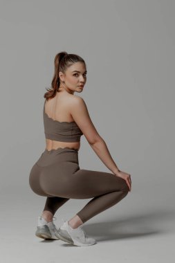 Uzun saçlı, geri çekilen bir kadın, atletik vücudunu sergileyen bir çömelme yeriyle meşgul. Eşleşen kahverengi bir giysi giyiyor ve gri arka planlı bir minimalist stüdyo ortamında..