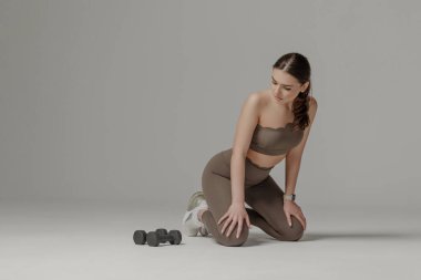 Atletik kıyafetli bir kadın düz bir yüzey üzerinde diz çöküp dambıl egzersizine odaklanıyor. Gri arka plan onun kararlılığını ve zindelik yolculuğunu vurguluyor..