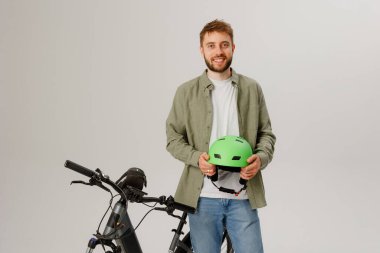 Genç bir adam yeşil bir kask tutarken gülümsüyor. Kapalı bir alanda elektrikli bir bisikletin yanında duruyor, gezintiye çıkmaya hazır. Giysileri rahat ve rahat..