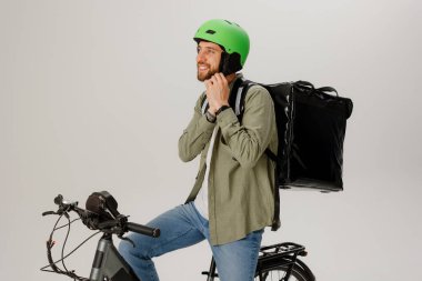 Bir teslimatçı bisikletle otururken kaskını ayarlar. Vardiyasına başlamaya hazır. Gülümsüyor ve sıradan bir kıyafet giyiyor. Sırt çantasıyla eşyaları taşımaya hazır..