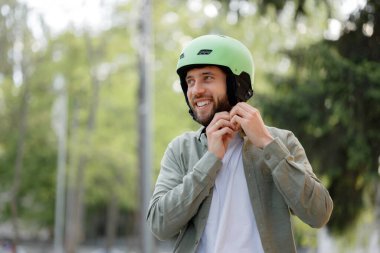 Neşeli bir suratı olan bir adam yeşil miğferini ağaçlarla çevrili bir parkta ayarlar. Sıradan giyinmiş, bisiklet ya da paten gibi eğlenceli bir açık hava aktivitesine hazır..