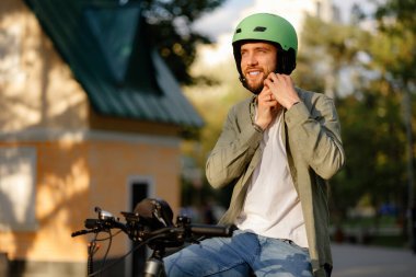 Bir adam güneşli bir parkta bisiklette otururken yeşil kaskını ayarlar. Bölgeyi çevreleyen ağaçlar ve arka planda görünen bir bina var. Bisiklet sürmek için güzel bir gün..