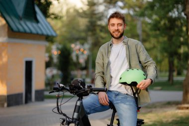Genç bir adam parkta bisikletinin üzerinde oturuyor, günlük kıyafetler giyiyor ve gülümsüyor. Bir elinde yeşil bir kask tutuyor, yeşil ve yumuşak güneş ışığıyla çevrili..