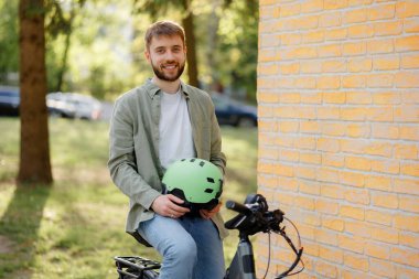 Sakallı genç bir adam elektrikli bisikletin üzerinde oturuyor, elinde yeşil bir kask var. Dışarıda ağaçlar ve güneş ışığıyla çevrili bir parkta, rahat ve mutlu görünüyor..