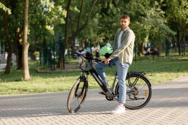 Genç bir adam yeşilliklerle çevrili canlı bir parkta elektrikli bisiklet sürüyor. Yeşil bir miğfer tutuyor ve gülümsüyor, güneşli bir günde dışarıda eğleniyor..