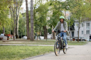Bir adam canlı bir parkta ağaçlarla kaplı bir dağ bisikletine biniyor, güneşli bir günde güzel havanın tadını çıkarıyor. İnsanlar arka planda çeşitli etkinliklerde görülebilir.