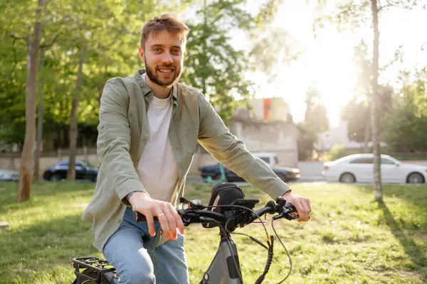 Yeşillik dolu bir parkta bisikletinin üzerinde oturan genç bir adam gülümsüyor. Güneş ışıl ışıl parlıyor ve açık hava aktiviteleri için sıcak ve davetkar bir atmosfer yaratıyor..