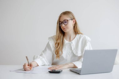 İşine odaklanmış genç bir kadın bir masa başında dizüstü bilgisayar ve hesap makinesiyle oturuyor. Gündüz vakti etrafı temiz ve parlak çalışma alanlarıyla çevrili kağıtlara notlar yazıyor..