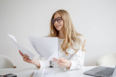 Genç bir kadın temiz bir masada oturuyor, birden fazla belgeyi incelerken gülümsüyor. Çalışma alanı parlak ve minimalistik, yakınında bir dizüstü bilgisayar ve hesap makinesi var..