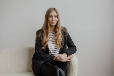 Genç bir kadın açık bir koltukta rahat bir şekilde oturur, çizgili bir gömleğin üzerine şık bir siyah ceket giyer. Uzun, dalgalı saçları minimalist arkaplanı tamamlıyor, sakin bir ifade sergiliyor..