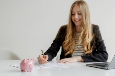 Uzun saçlı bir kadın bir masada mali belgeler yazmakla meşgul. Bilgisayarının yanında pembe bir kumbara duruyor. Modern bir ofiste tasarruf ve bütçeyi sembolize ediyor..