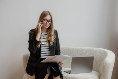 Resmi giyinmiş genç bir kadın şık bir kanepede oturur ve bir telefon görüşmesi yapar. Bir elinde bir dizüstü bilgisayara bakıp üretken bir çalışma ortamı sunuyor..