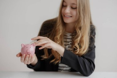 Uzun saçlı bir kadın pembe bir kumbara tutarken siyah ceket ve çizgili gömlek giyer. Parlak kapalı alan neşeli bir atmosferi vurguluyor..