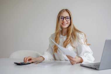 Masada otururken gülümseyen genç bir bayanın olduğu parlak bir ofis ortamı. Bir elinde kağıtları tutuyor, diğer elinde ise üretkenlik ve odaklanma gösteren bir hesap makinesi kullanıyor..