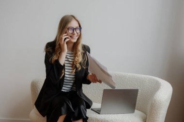Siyah ceketli ve çizgili gömlekli genç bir kadın telefonda konuşurken gülümsüyor. Bir elinde dokümanları tutuyor ve bir dizüstü bilgisayarın yanına şık bir sandalyeye oturuyor, yoğun bir iş günü olduğunu gösteriyor..