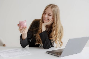 Genç bir kadın, elinde pembe bir kumbara tutarken masasında vakit geçirmekten zevk alıyor. Dizüstü bilgisayarı ve mali belgeleri mali durumunu parlak bir ortamda planladığını gösteriyor..