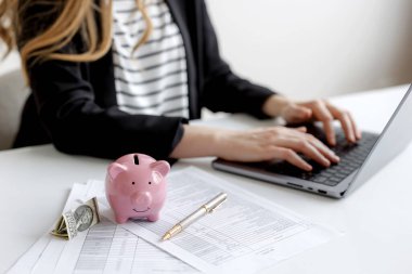 Bir kadın, temiz bir masadaki mali belgeleri incelerken dizüstü bilgisayarda yazar. Pembe bir domuz kumbarası ve birkaç dolarlık banknotlar da mevcut. Çalışma alanına kişisel bir dokunuş da eklendi..