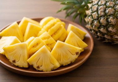 Taze, soyulmuş ve dilimlenmiş ananas parçalarıyla dolu ahşap bir tabak, arka planda bütün bir ananas, rustik ahşap bir masa üzerine kurulmuş..