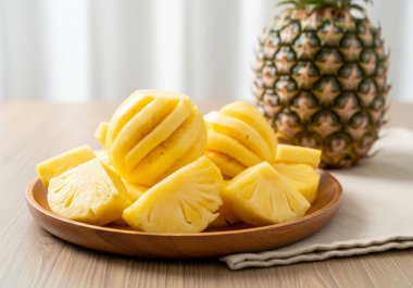 Taze, soyulmuş ve dilimlenmiş ananas parçalarıyla dolu ahşap bir tabak, arka planda bütün bir ananas, rustik ahşap bir masa üzerine kurulmuş..
