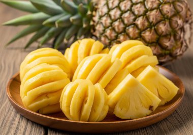 Taze, soyulmuş ve dilimlenmiş ananas parçalarıyla dolu ahşap bir tabak, arka planda bütün bir ananas, rustik ahşap bir masa üzerine kurulmuş..