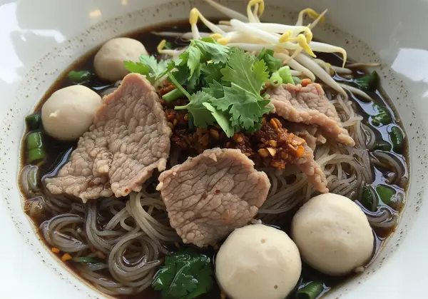 Dilimlenmiş domuz eti, et, köfte, fasulye filizi ve taze otlar ve çıtır sarımsak ile süslenmiş, zengin, koyu renk bir çorbanın içindeki sabah zaferiyle dolu canlı bir kase otantik Tayland eriştesi (Guay Teow Rua)..