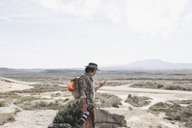 Şapkalı ve sırt çantalı bir gezgin elinde bir kamerayla akıllı telefona danışıyor..