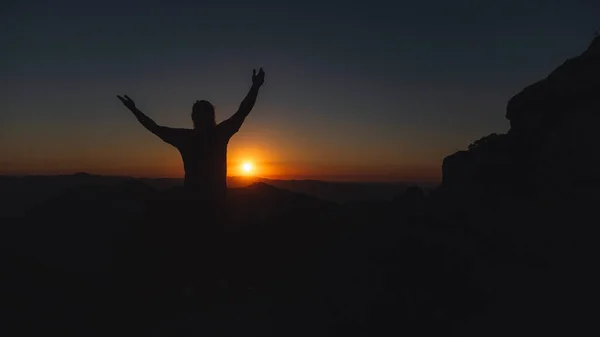 A man standing with his arms open on a mountain as the sun sets.