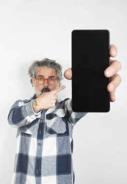 Beyaz bir arkaplanda işaret parmağıyla işaret eden boş bir ekranlı cep telefonu tutan bir adamın yakın çekimi.