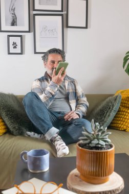 Evde akıllı telefon asistanı kullanarak koltukta dinlenen olgun bir adam.