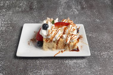 Krem şantili, yabanmersinli, çilekli, karamel ve tarçınlı beyaz tabakta oturan bir dilim pasta.