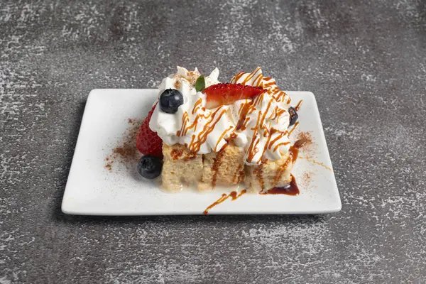 Krem şantili, yabanmersinli, çilekli, karamel ve tarçınlı beyaz tabakta oturan bir dilim pasta.