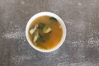 Japon mutfağını temsil eden dokulu gri bir arka planda beyaz bir kasede Wakame yosunlu, mantarlı ve yeşil soğanlı Miso çorbası servis ediliyor.