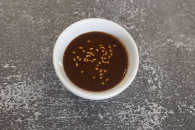 Üzerinde susam tohumları serpiştirilmiş, gri desenli yüzeyde duran küçük beyaz seramik kâse.