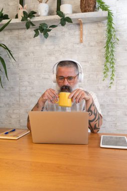 Kulaklıklı, kahve içen ve bilgisayarını kullanarak uzaktan çalışan kıdemli bir adam.