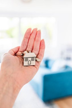 hand holding house shaped keyring with key in front of living room bokeh, dream home and home ownership concept (the key is an old key that doesn't open anything) clipart