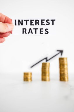 Elele tutuşan Faiz Oranları, arka planda ok olan ve aşırı fiyatlandırma veya spekülasyon simgesi olarak büyüyen madeni para yığınları önünde metin tutar