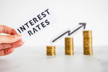 Elele tutuşan Faiz Oranları, arka planda ok olan ve aşırı fiyatlandırma veya spekülasyon simgesi olarak büyüyen madeni para yığınları önünde metin tutar