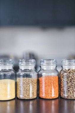 İçinde kinoa mercimek, karabuğday ve arpanın da bulunduğu farklı türde tahıl ve baklagillerin bulunduğu açık kiler kavanozları mutfak tezgahı, basit malzeme konsepti.