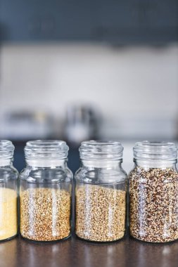 İçinde kinoa mercimek, karabuğday ve arpanın da bulunduğu farklı türde tahıl ve baklagillerin bulunduğu açık kiler kavanozları mutfak tezgahı, basit malzeme konsepti.