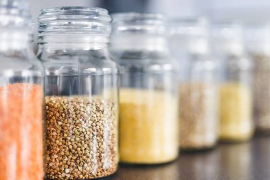 İçinde kinoa mercimek, karabuğday ve arpanın da bulunduğu farklı türde tahıl ve baklagillerin bulunduğu açık kiler kavanozları mutfak tezgahı, basit malzeme konsepti.